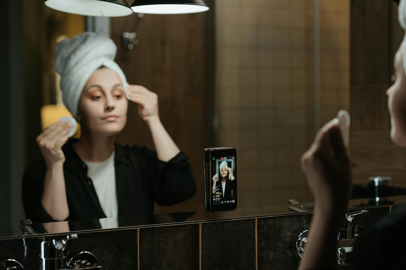 Photo by cottonbro studio A woman applies skincare products while filming herself in a bathroom mirror with a smartphone.