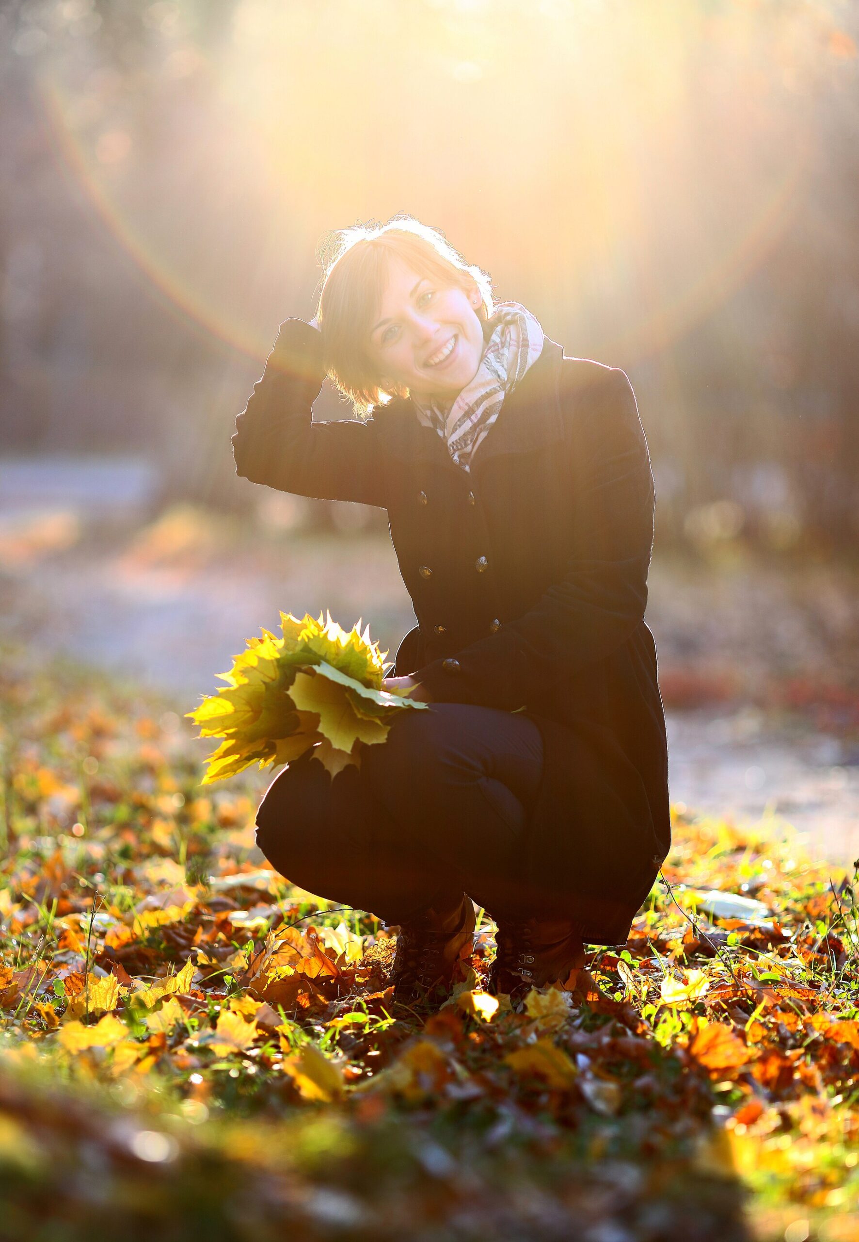 girlook Femme accroupie dans les feuilles d'automne, un bouquet de feuilles à la main, éclairée par le soleil bas